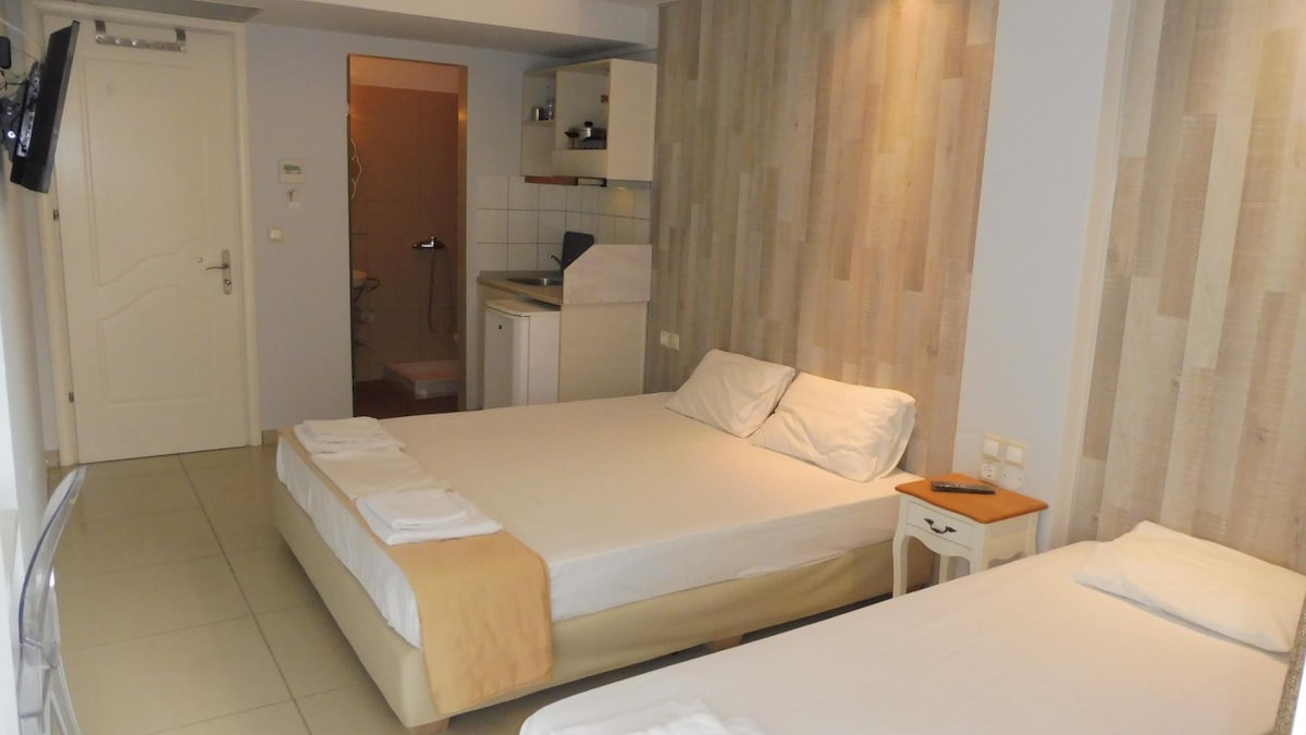 A well-lit room features two spacious beds with white linens arranged neatly. A small bedside table holds a remote control. The adjoining area is equipped with a kitchenette, and light-colored wall panels enhance the bright, open feel of the space.