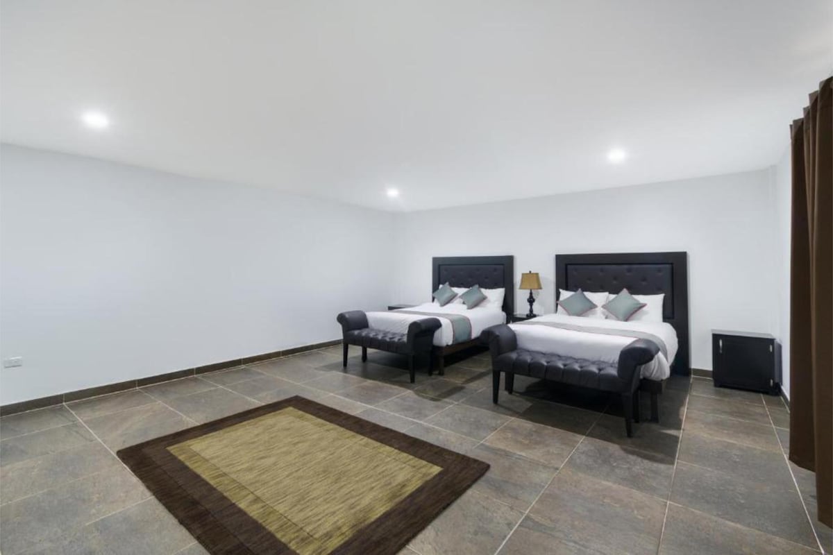 A spacious room features two large beds with dark upholstered headboards, complemented by neutral bedding accents. A plush area rug sits on a tiled floor, while a side table with a lamp is placed beside one of the beds. Soft lighting illuminates the simple yet modern design.