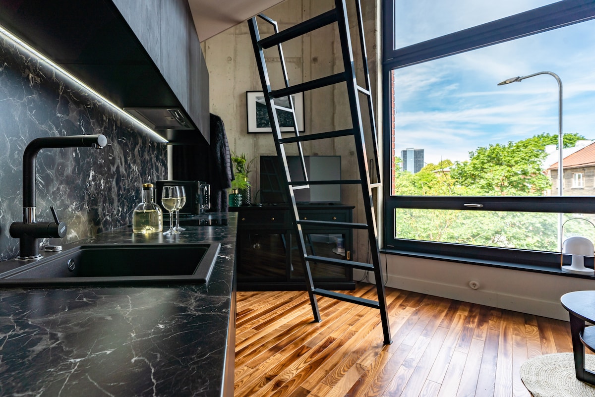 A modern kitchenette features dark cabinetry and a sleek black countertop. A ladder leads to the upper sleeping area. Large windows allow natural light to fill the space, with views of greenery outside and a hint of city buildings in the distance. Warm wooden flooring complements the design.