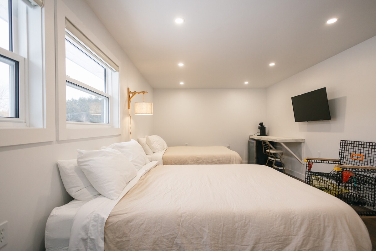 A bright room features two spacious beds dressed in neutral bedding. Natural light enters through two windows, illuminating the space. A small work area with a bar stool and a coffee maker is positioned against the wall. A flat-screen TV is mounted nearby.