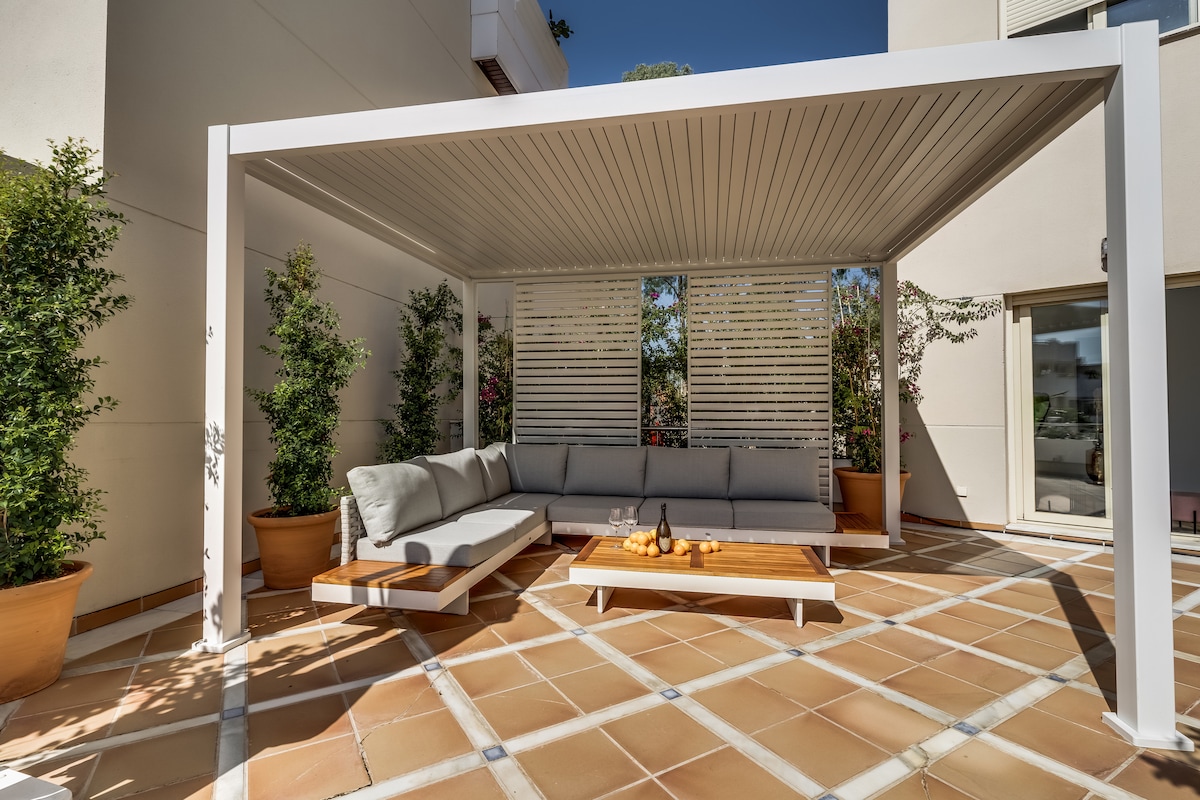 A covered outdoor seating area features a spacious sectional sofa arranged around a wooden coffee table. Potted plants add greenery to the sunlit terrace. The design creates a comfortable space for relaxation, with ample shade offered by the pergola overhead.
