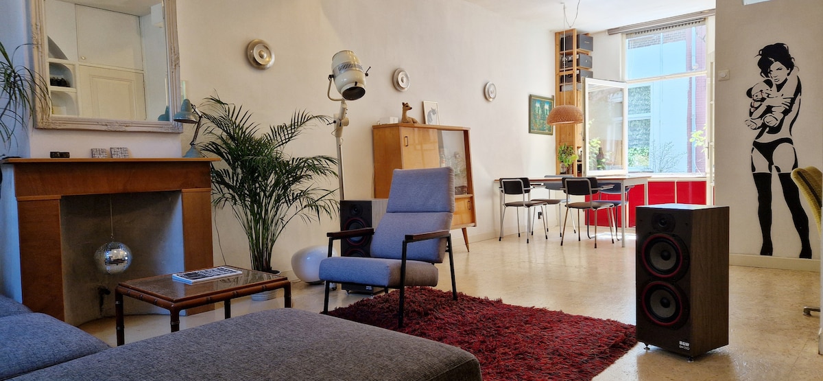 A spacious living area features a comfortable gray sofa, an accent chair, and a side table. Natural light enters through large windows, creating a bright atmosphere. A dining area is visible in the background, accented by a decorative wall art piece and greenery.