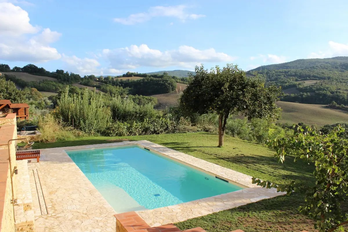 A heated saltwater swimming pool is surrounded by a stone deck, set against a backdrop of rolling Tuscan hills. A single tree provides shade, and the landscape features lush greenery and distant hills under a bright blue sky with scattered clouds.
