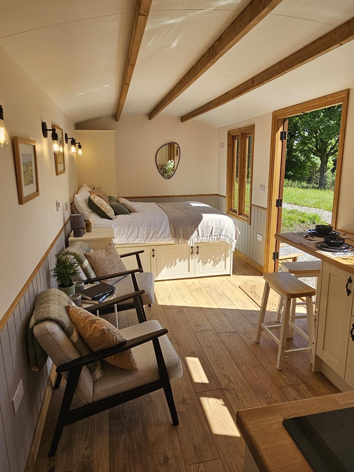 An inviting interior features a spacious King-Sized bed with soft linens and natural light streaming through wooden-framed windows. A cozy seating area with two chairs enhances the comfort, while wooden beams accentuate the ceiling, creating a welcoming atmosphere.