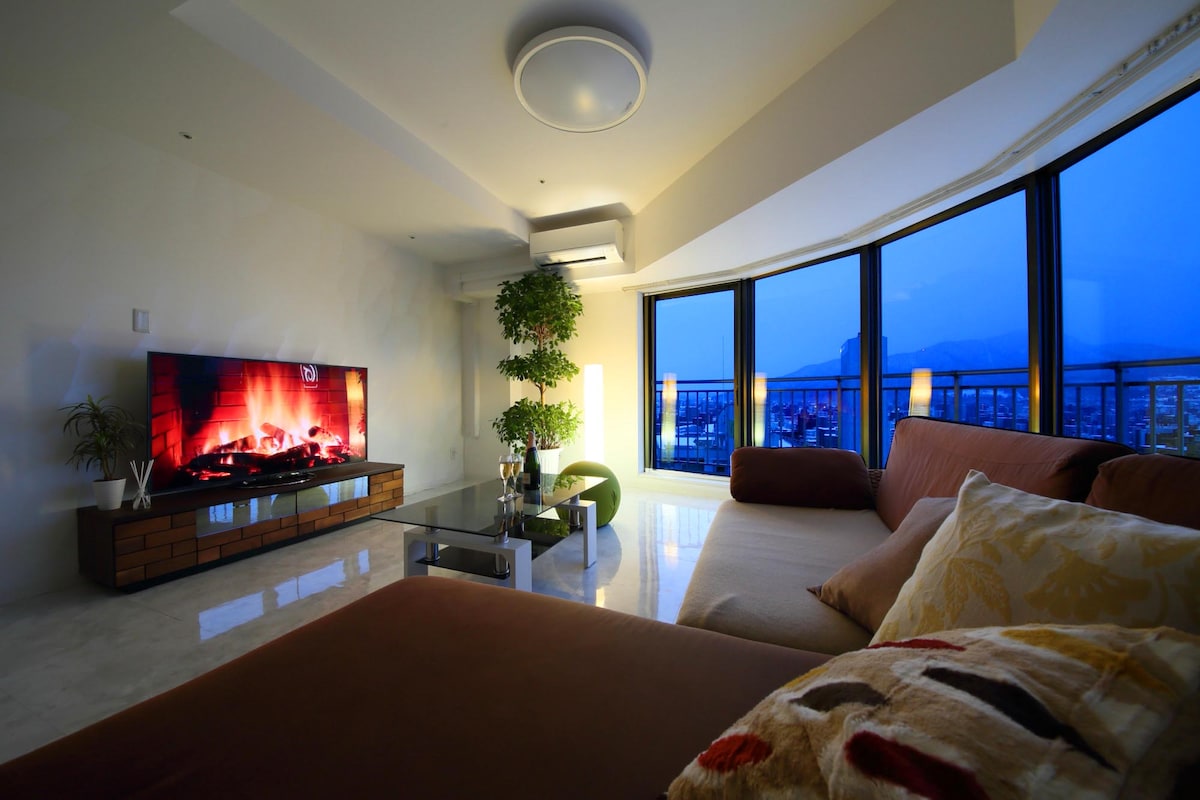 A spacious living area features floor-to-ceiling windows that offer city views during twilight. A modern television displays a calming fireplace scene, while a large sofa adorned with decorative pillows invites relaxation. Indoor plants add a touch of greenery to the decor.
