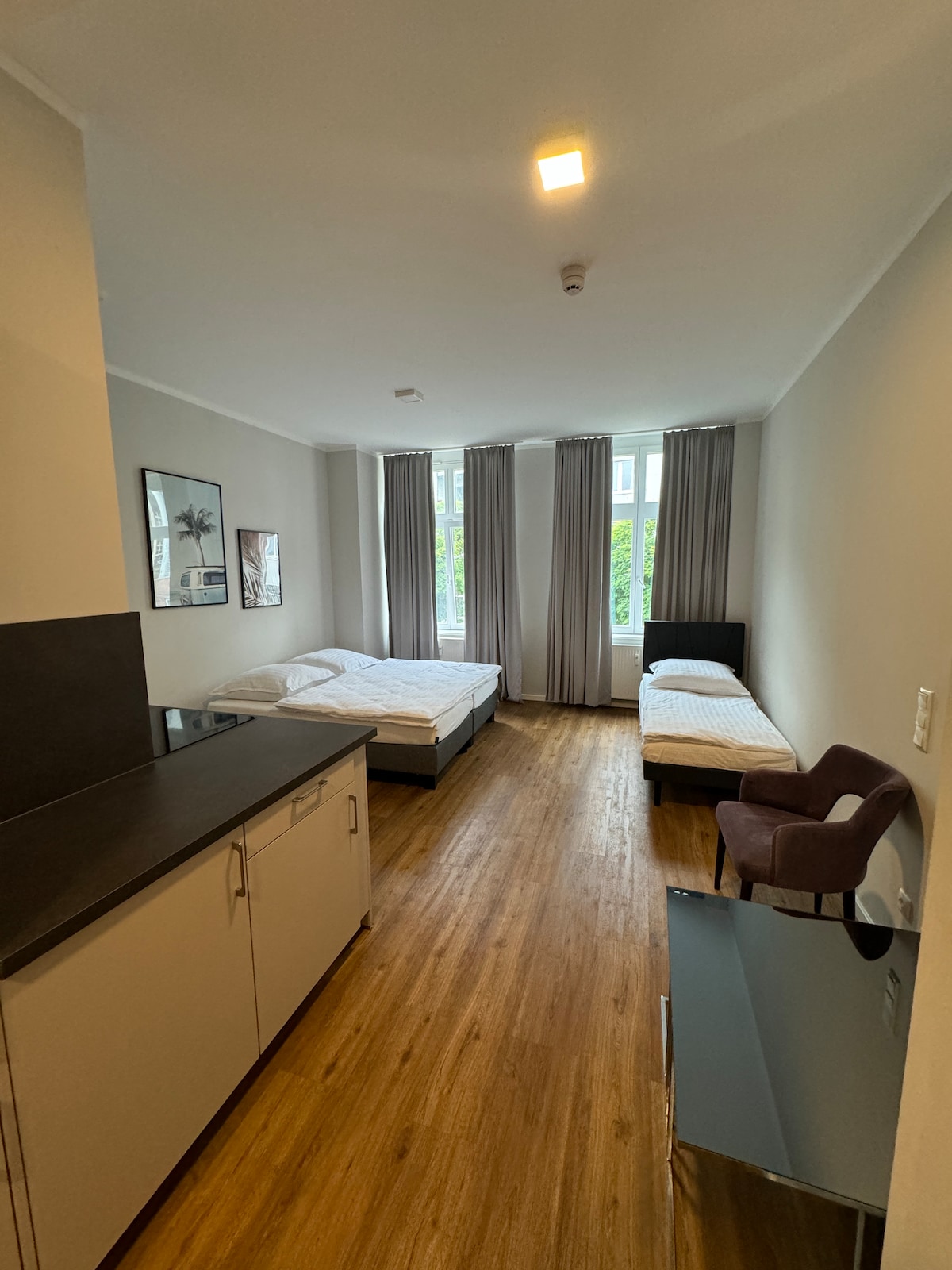 The room features two beds arranged for comfortable sleeping, with a spacious layout. Tall windows provide natural light, framed by floor-length curtains. A small chair and a compact table are included, along with a kitchenette area equipped for basic meal preparation.