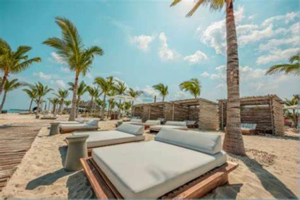 Lounge chairs with white cushions are positioned on sandy terrain under swaying palm trees. Wooden structures provide shaded areas in the background, contrasting with the clear blue sky and scattered clouds above.