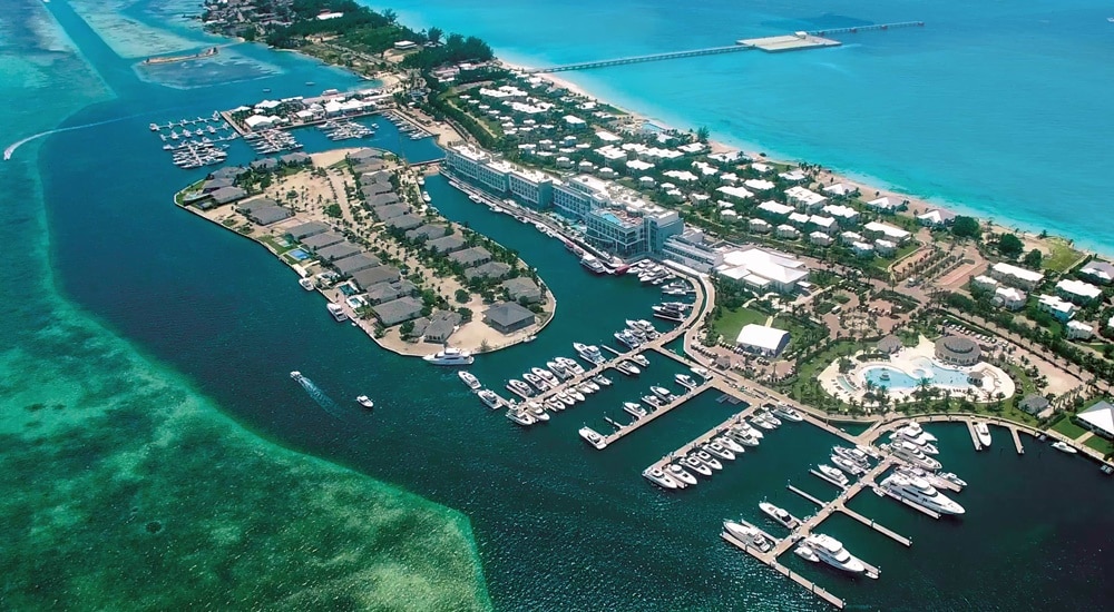 An aerial view showcases the marina and nearby amenities of Bimini Bay. Expansive turquoise waters surround the vibrant green landscape, dotted with boats and yachts. The resort features well-maintained structures alongside a picturesque harbor, creating a serene coastal atmosphere.