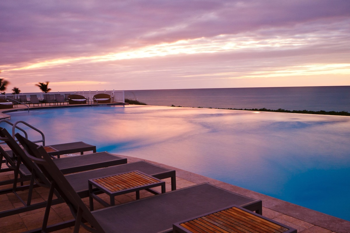 An ocean-view infinity pool reflects soft pastel colors from a sunset sky. Comfortable lounge chairs with wooden tabletops are arranged around the pool, inviting relaxation while taking in the scenic backdrop of the serene sea.