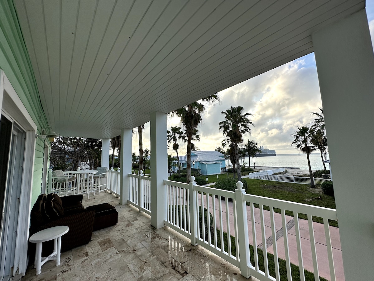 A spacious balcony is lined with white railings, featuring comfortable seating arrangements. A view of palm trees and the ocean horizon is visible, along with a pathway leading to the beach. The sky showcases soft clouds, adding to the tranquil atmosphere.