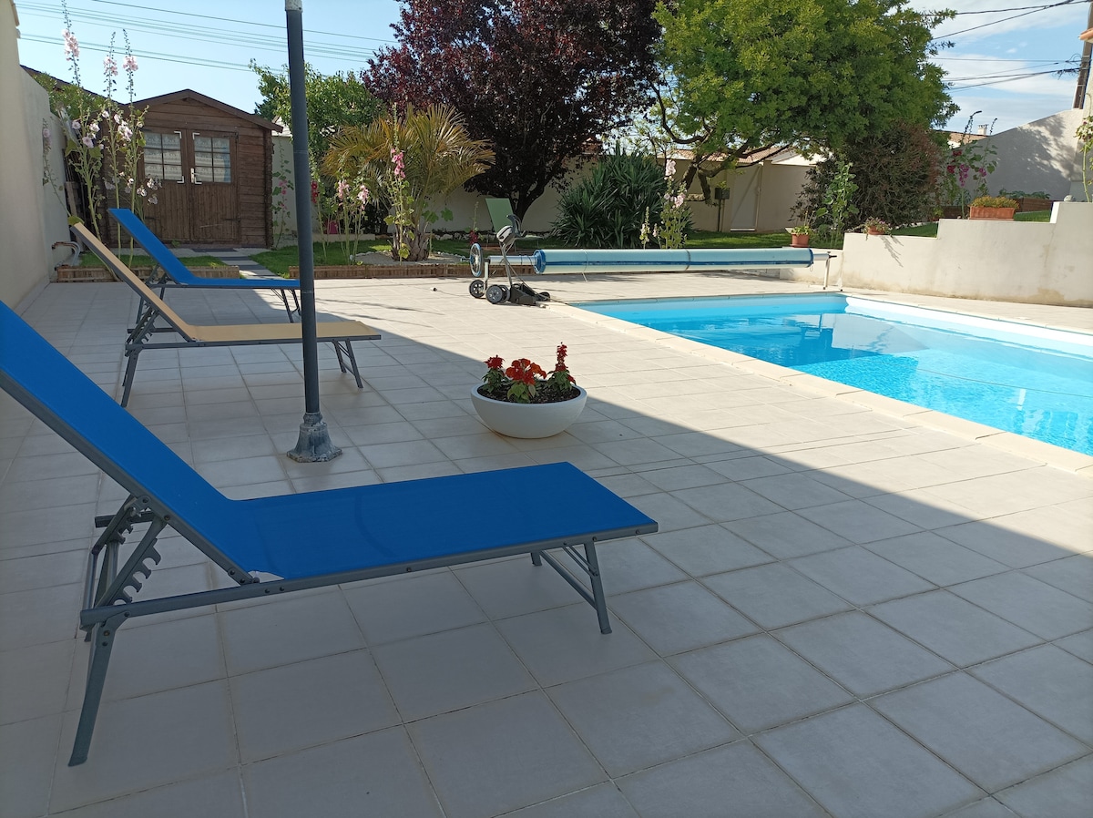 A private outdoor space features a sparkling turquoise pool surrounded by tiled flooring. Sun loungers are positioned nearby, and a planter filled with vibrant flowers adds a touch of color. A wooden shed and green foliage are visible in the background, enhancing the serene atmosphere.