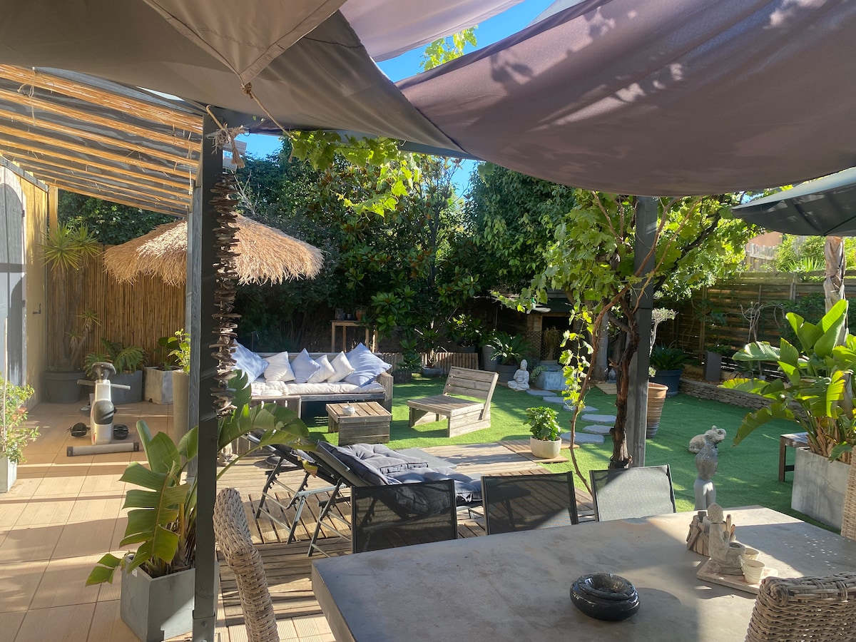 The outdoor space features a well-maintained garden with various plants and trees. A shaded area with lounge seating is visible, along with a dining table beneath a canopy. Decorative elements, including sculptures, enhance the tranquil ambiance of the surroundings.