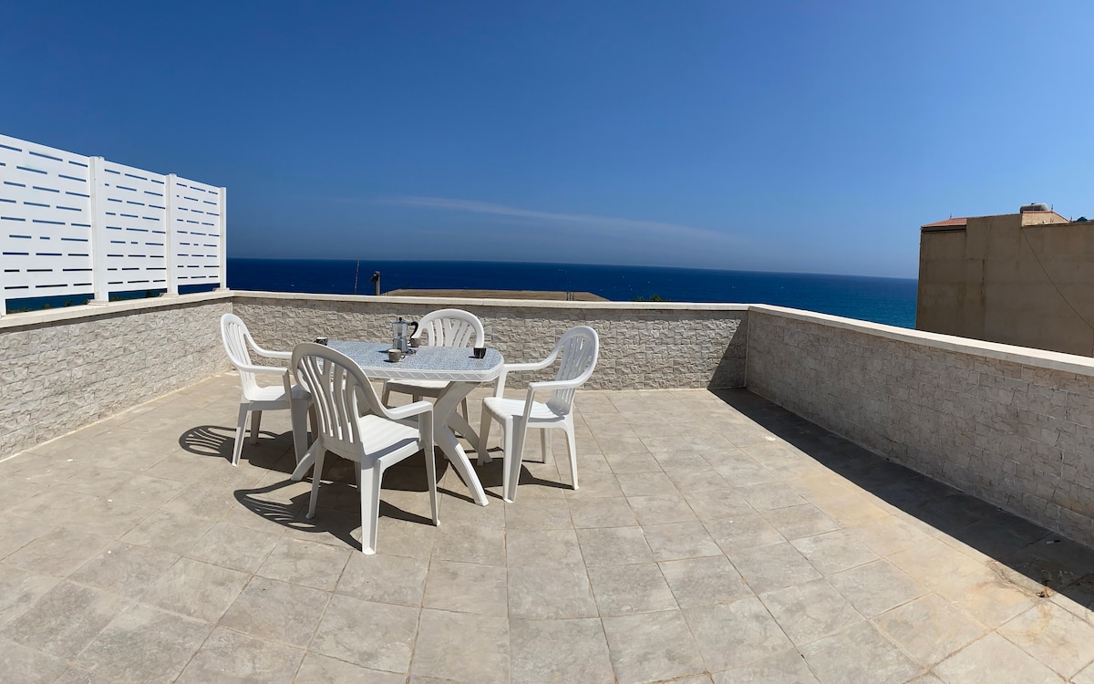 A spacious terrace is presented, featuring a round table surrounded by four white chairs. A clear view of the sea stretches into the horizon, complemented by the bright blue sky. The stone wall adds a subtle backdrop to the inviting outdoor space.