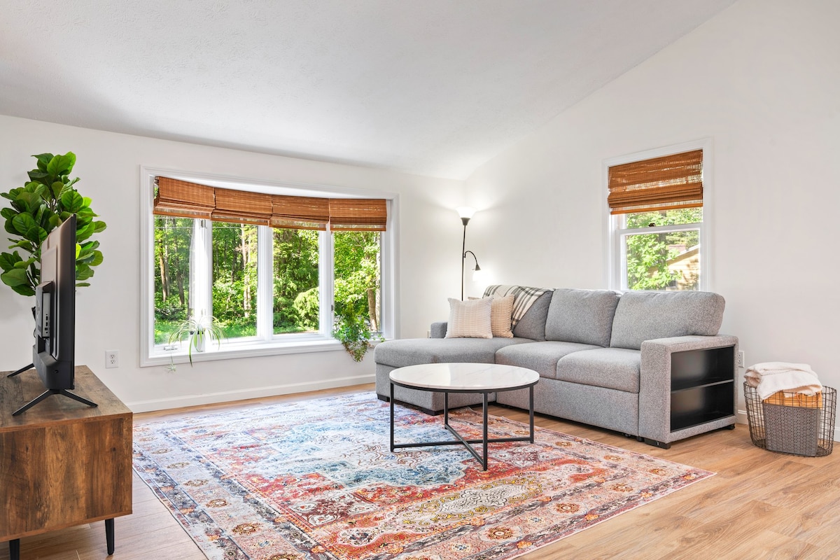 A light-filled living area features a comfortable grey sectional sofa and a round coffee table. A neutral-toned area rug lies beneath, while large windows with bamboo shades overlook a green outdoor space, creating a connection with nature. A TV console is positioned near the television.