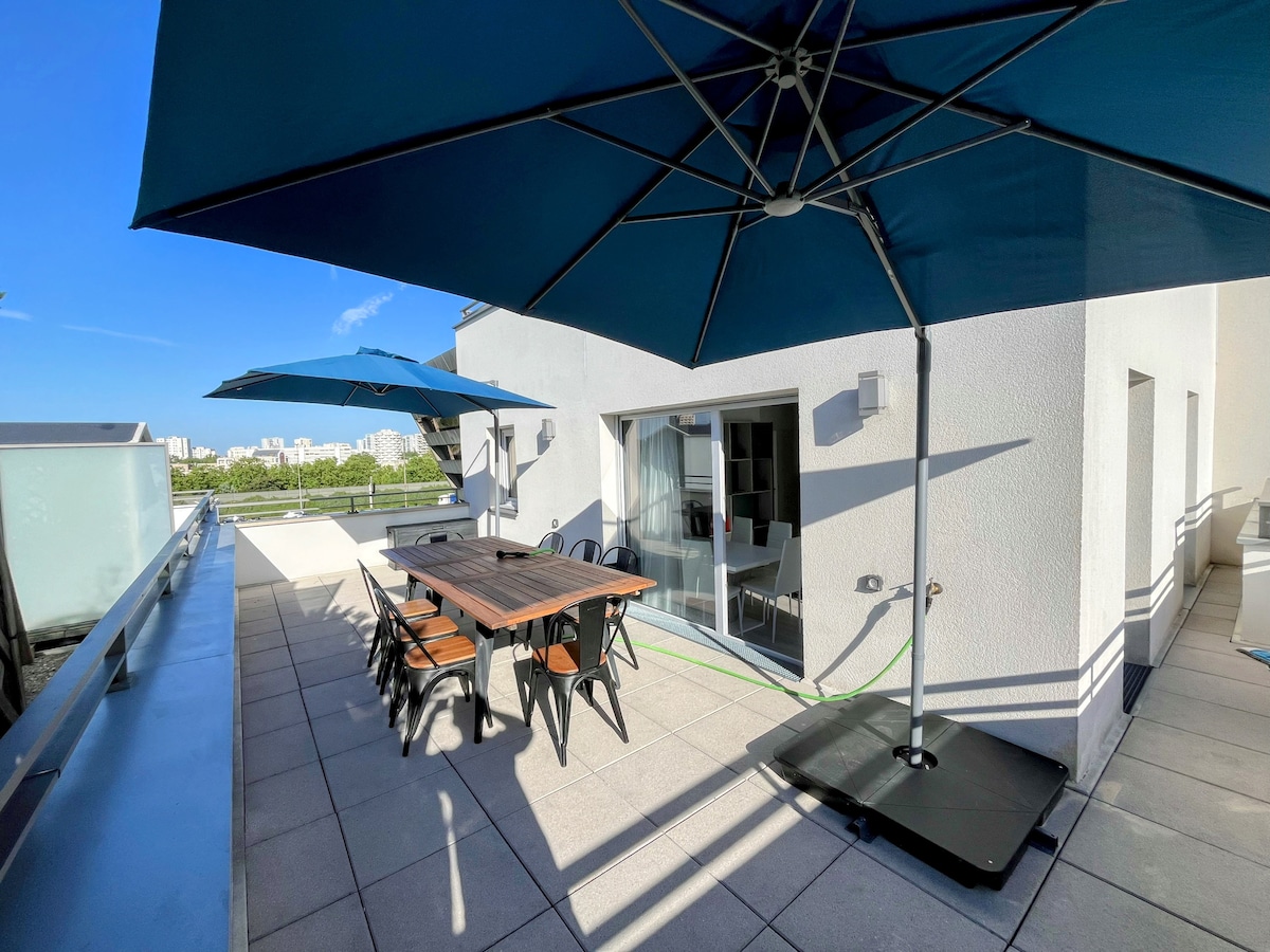 A spacious terrace features a wooden dining table surrounded by six chairs, shaded by two large umbrellas. The area is paved with light-colored tiles, and sliding glass doors lead into the indoor space. A scenic view of the surroundings is visible in the background.