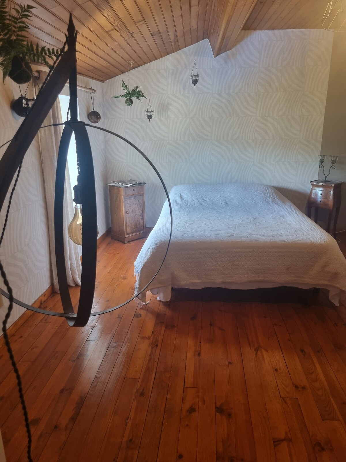 A cozy bedroom features a spacious bed covered with a light quilt. The wooden floor adds warmth, while the textured wallpaper complements the natural light filtering through a window. Decorative plants hang from the ceiling, enhancing the room's inviting atmosphere.