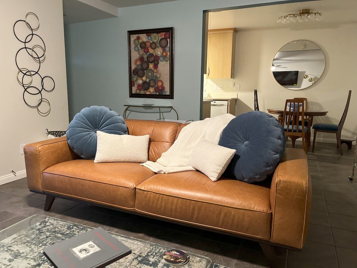 A cozy seating area is featured with a leather sofa adorned with large blue pillows and white accents. A textured throw is draped casually over the arm. In the background, a dining area with a round mirror and wood table can be seen, enhancing the space's open layout.