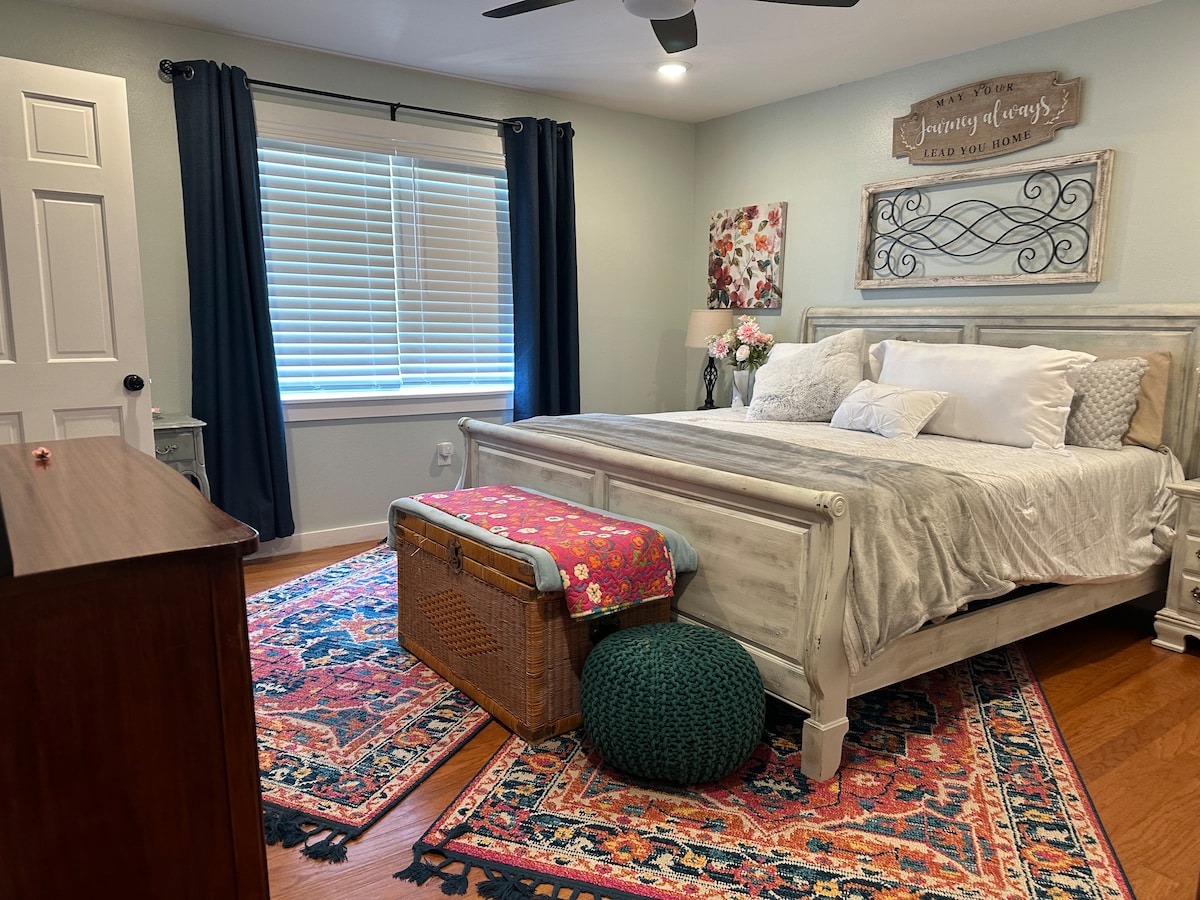 A cozy bedroom features a spacious bed with decorative pillows and a woven throw at the foot. Warm wood floors are enhanced by a patterned area rug. A dresser is positioned nearby, and a window with blinds allows natural light to enter, framed by dark drapes.