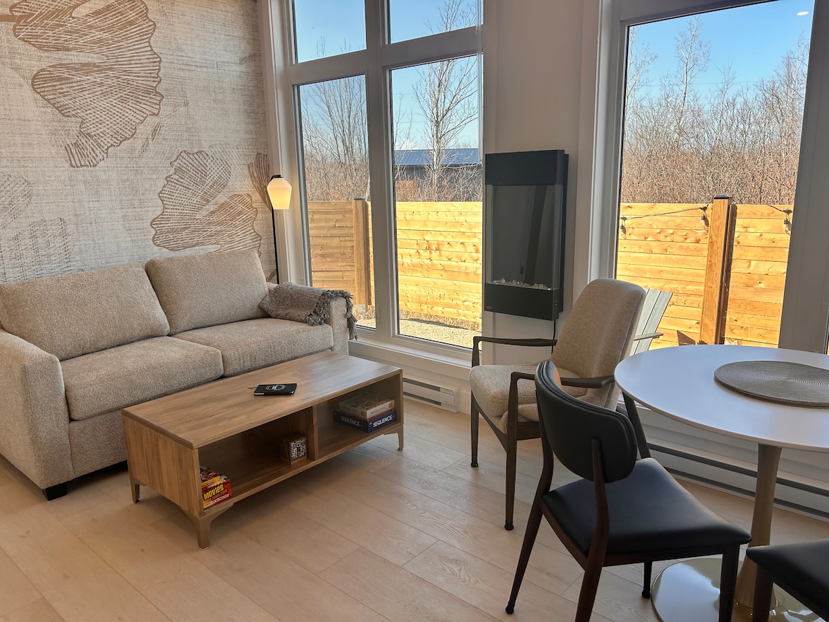 A modern living space features a light-colored sofa, a wooden coffee table with a few books, and a sleek chair beside a round dining table. Large windows allow natural light to illuminate the room, with a view of a wooden fence and greenery outside.