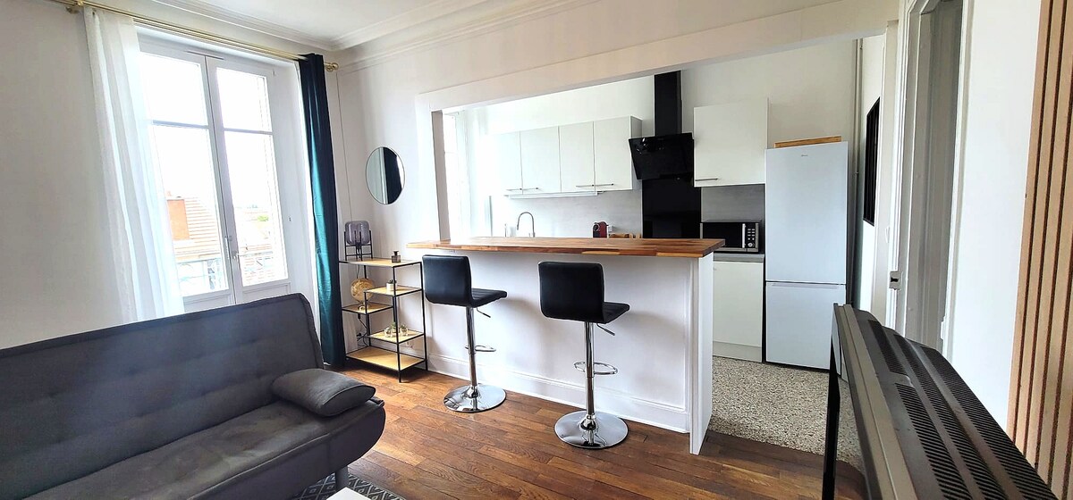 A modern open-plan layout is displayed, featuring a sleek kitchen bar with two black stools. A comfortable sofa is positioned nearby, while the kitchen includes white cabinets and essential appliances. Large windows add natural light, enhancing the airy feel of the space.