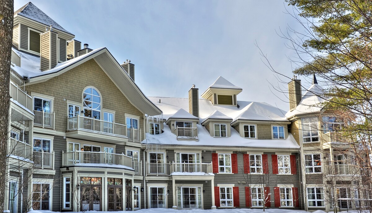 A multi-story building is set against a snowy backdrop, featuring a combination of balconies and distinctive architectural elements. The facade displays a mix of muted colors with large windows, allowing for ample natural light, while the roof is covered in snow.