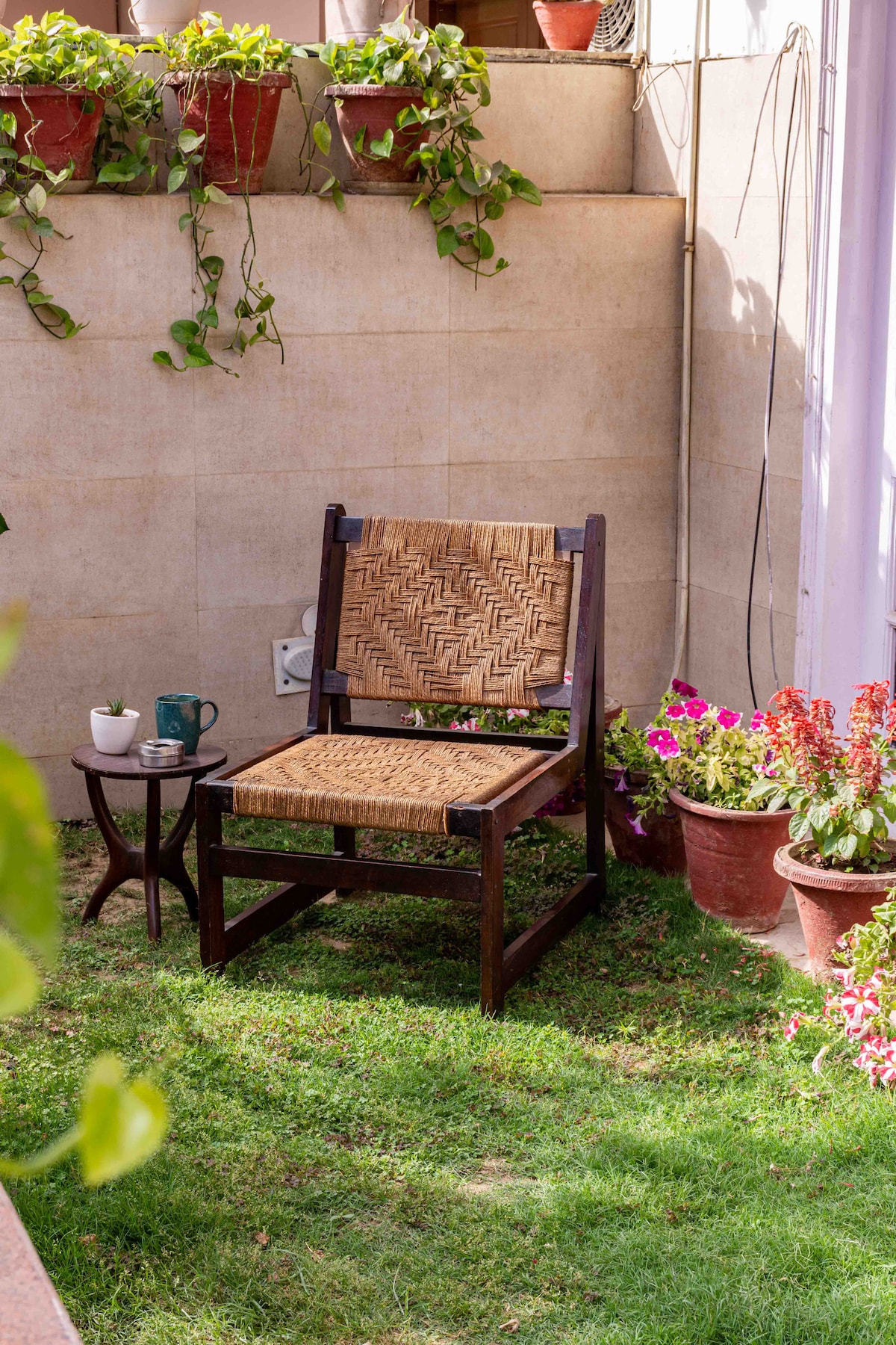 A wooden chair with a woven seat is positioned on the lush green lawn, accompanied by a small table. Potted plants with vibrant flowers and climbing greenery decorate the surrounding area, creating a serene outdoor corner for relaxation.