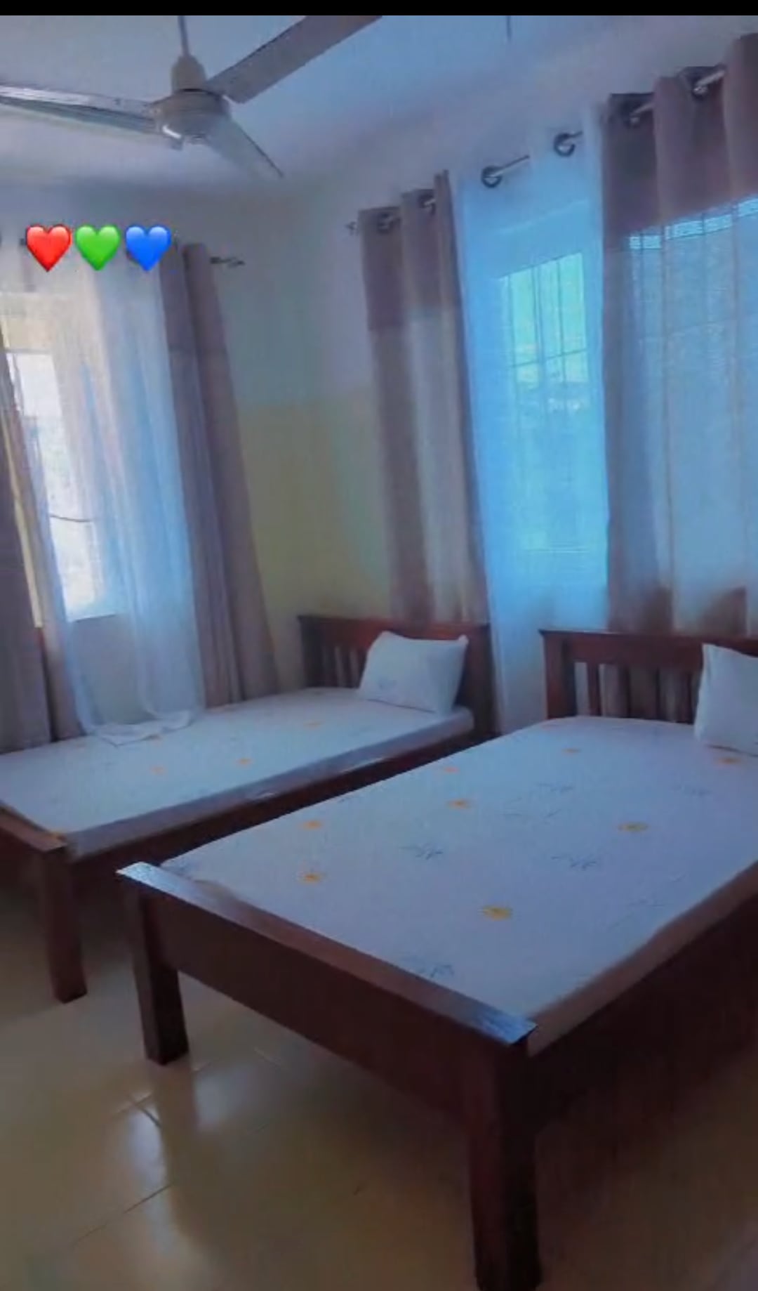 Two twin beds are positioned in a bright room, each accompanied by a white pillow. Natural light enters through large windows, enhanced by light curtains. The floors are tiled, and a ceiling fan is visible above, contributing to a comfortable ambiance.