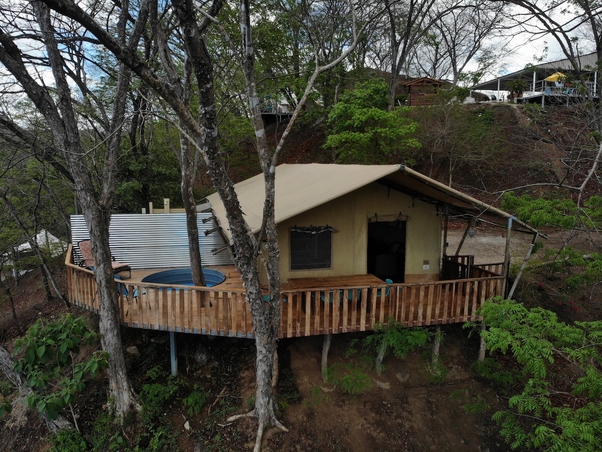 A unique cabin is nestled among trees, featuring a wooden deck that wraps around its exterior. The structure has a tent-like design with earthy tones, complemented by a small circular pool on the side. Surrounding nature adds to the tranquil setting.