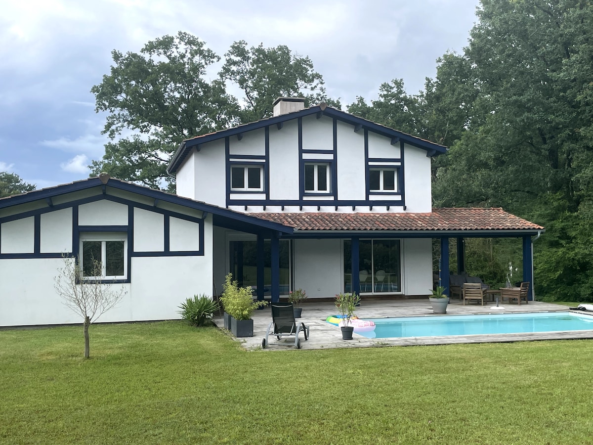 A two-story house with a white exterior accented by blue trim is set amidst greenery. A covered porch leads to a swimming pool surrounded by stone tiling and chairs. A lawn extends in front of the house, featuring a few potted plants and a small tree.