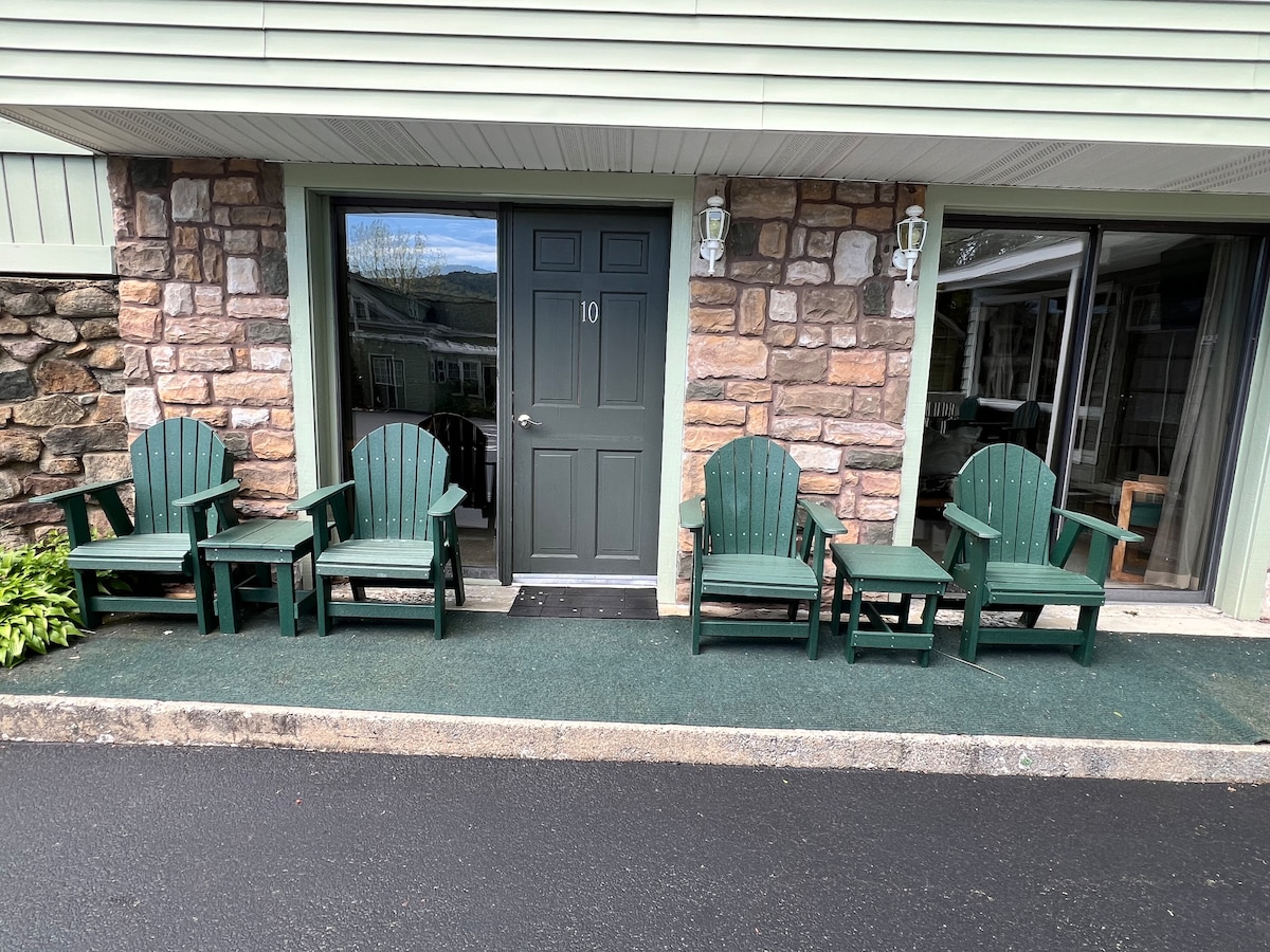 A welcoming entryway features a set of four green Adirondack chairs arranged beside a small side table, positioned in front of a green door framed by stone walls. The area is well-maintained with a carpeted entrance and a view of the surrounding nature.