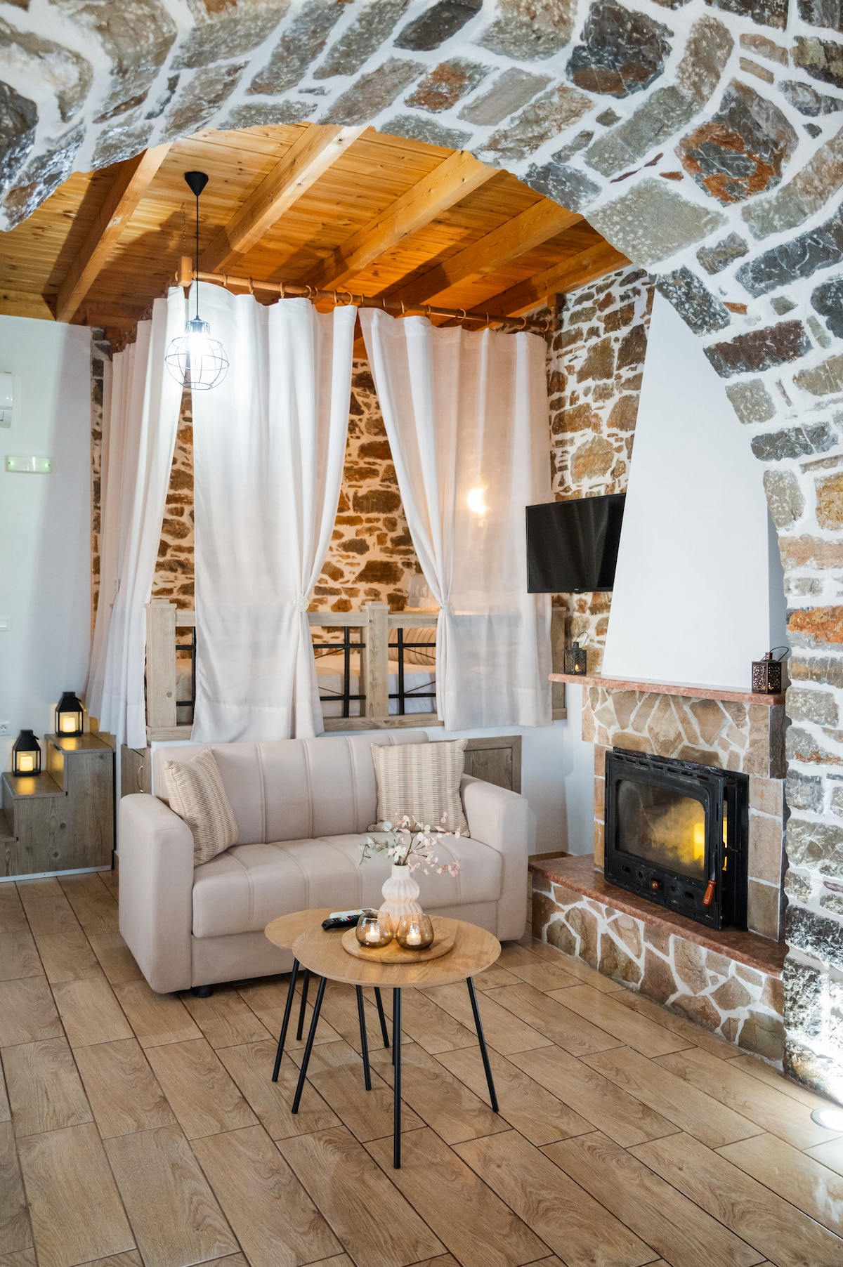 An inviting living area features a light-colored sofa paired with a small, round table. Exposed stone walls provide a rustic backdrop, while soft curtains frame the space. A warm fireplace is situated beside the seating area, complemented by ambient lighting from lanterns.