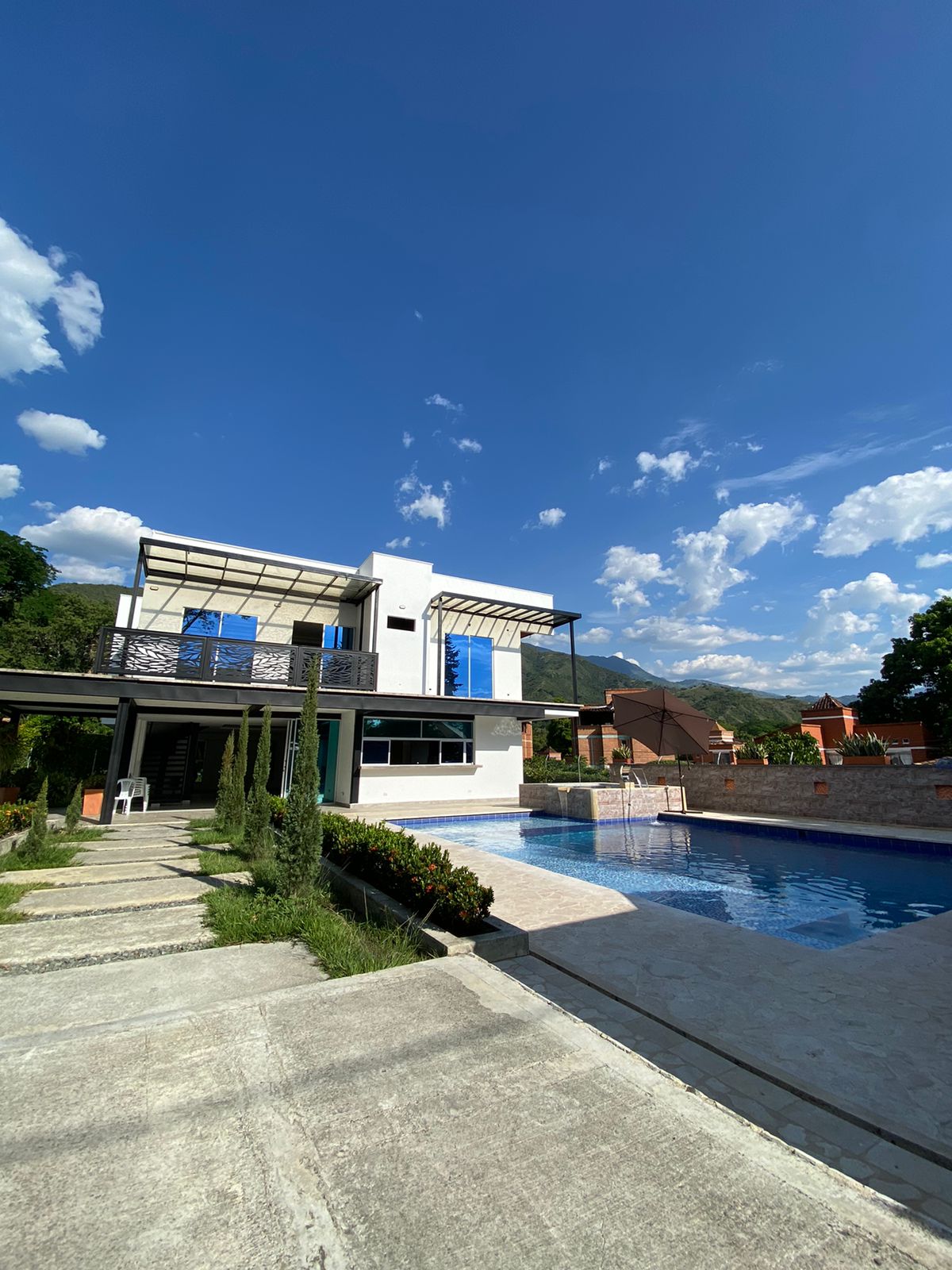 A modern multi-level villa is viewed from a landscaped path leading to a clear blue swimming pool. The building features large windows and a covered terrace, surrounded by green trees and mountains in the background under a partly cloudy sky.