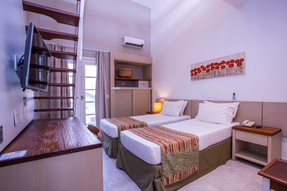 A well-appointed room features two spacious beds dressed in light linens, complemented by decorative throw blankets. A LED TV is mounted on the wall, and a staircase leads to an upper level. Natural light floods through the window, enhancing the neutral color palette.