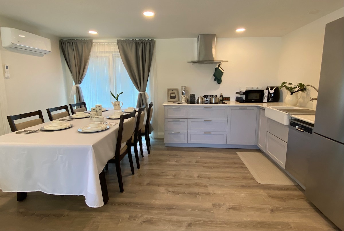 A bright kitchen area features a dining table set for six, with plates and utensils arranged neatly. Modern cabinetry and appliances are visible, including a fridge and microwave. Natural light streams through sheer curtains, creating an inviting space for meals and gatherings.