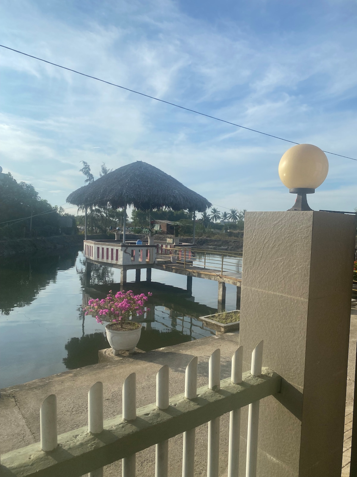 A scenic outdoor area features a thatched-roof structure on stilts above calm water, surrounded by lush greenery. A decorative pot with blooming flowers adds a colorful touch, while a lamp post stands at the edge of the pathway, enhancing the tranquil riverside setting.