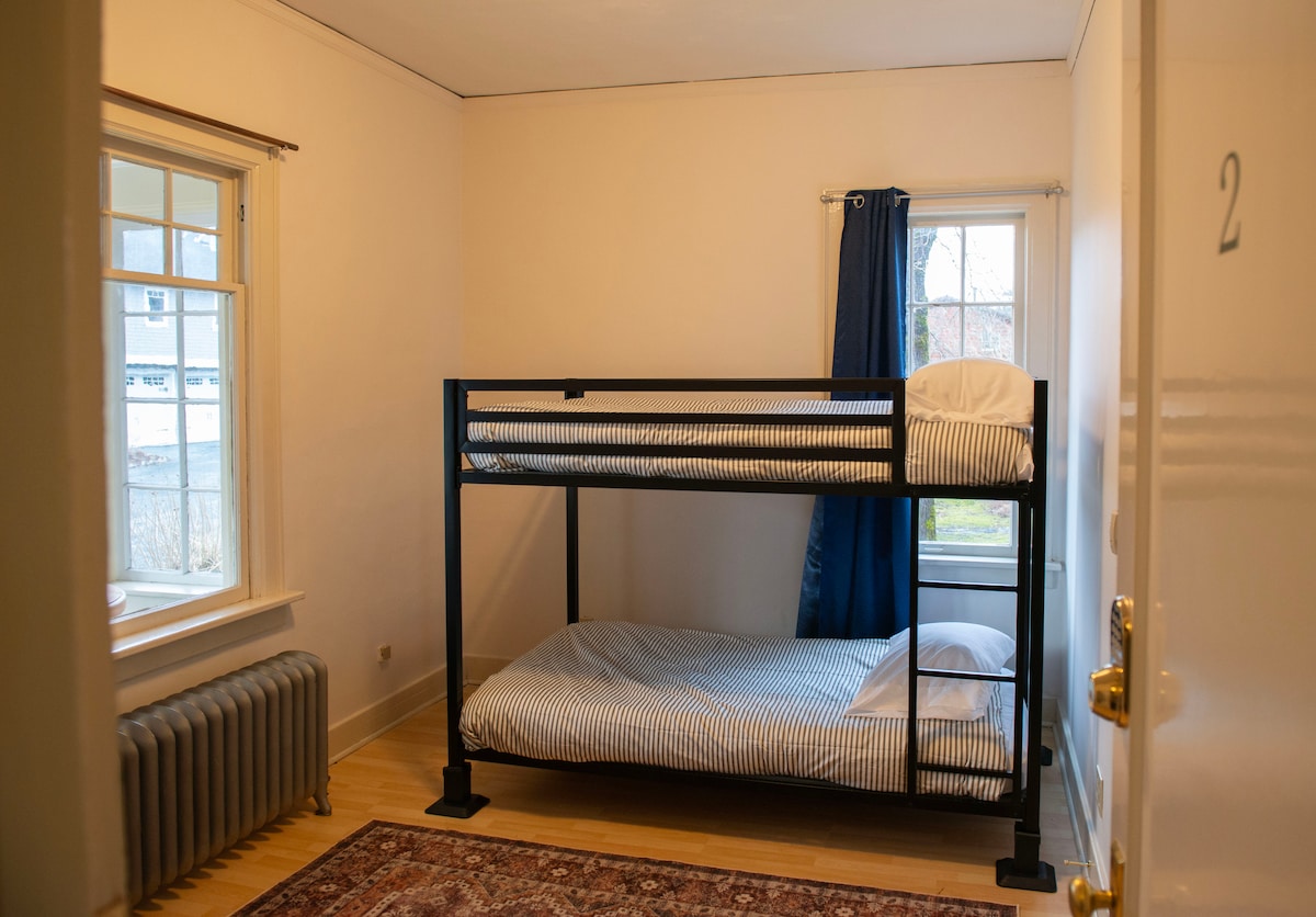A bright room features a metal bunk bed with striped bedding, providing sleeping space for two. Natural light enters through two windows adorned with blue curtains. A radiator adds warmth along the wall, while a simple rug adds a touch of comfort to the wooden floor.