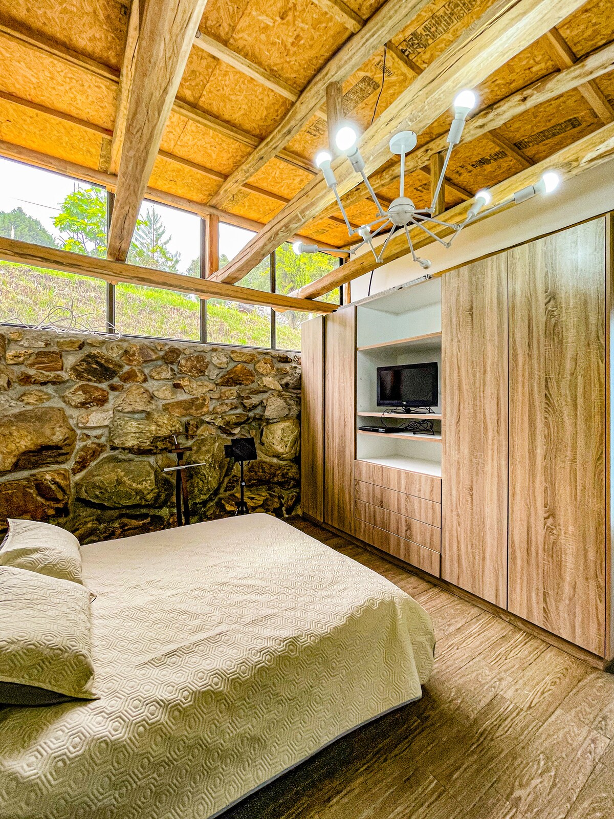 A serene bedroom features natural stone walls and a wood beamed ceiling. A spacious bed is adorned with a light-colored blanket. A modern entertainment unit is integrated into the cabinetry, with a television visible. Soft lighting enhances the calming ambiance of the room.