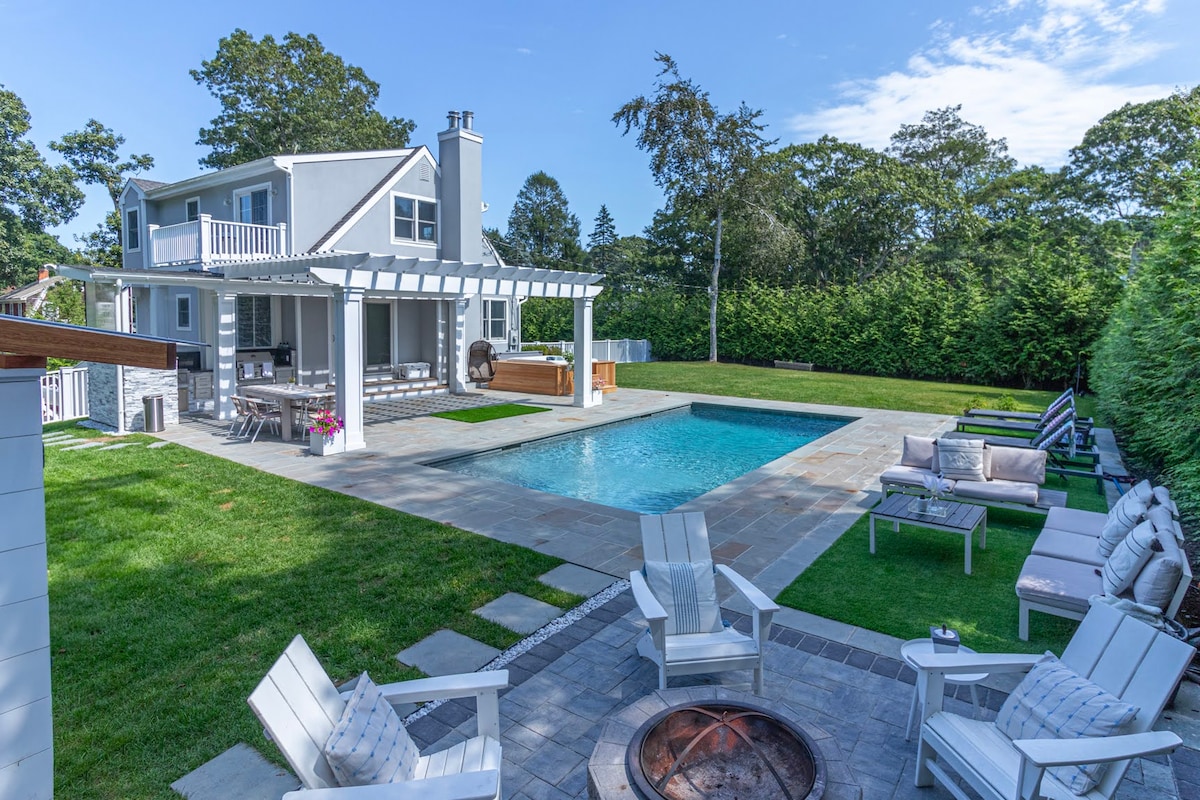 An outdoor area features a rectangular pool surrounded by stone patio. Comfortable seating is arranged near a fire pit, with a covered dining area visible. Lush greenery provides privacy, while a charming structure stands in the background, complete with a second-floor balcony.