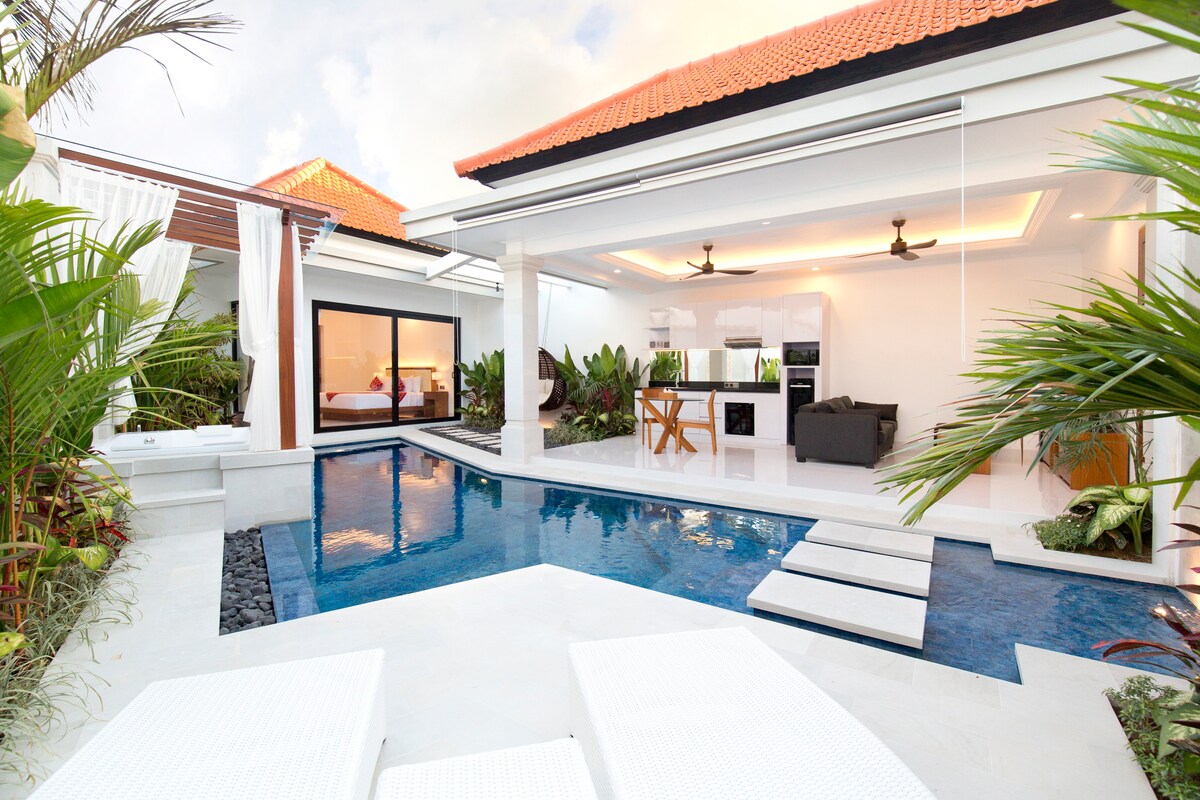 A serene outdoor space is highlighted by a private swimming pool, complemented by sunbeds and tropical plants. The living area, featuring an open layout with a comfortable sofa, dining table, and kitchenette, seamlessly connects to the pool area, creating a harmonious indoor-outdoor experience.