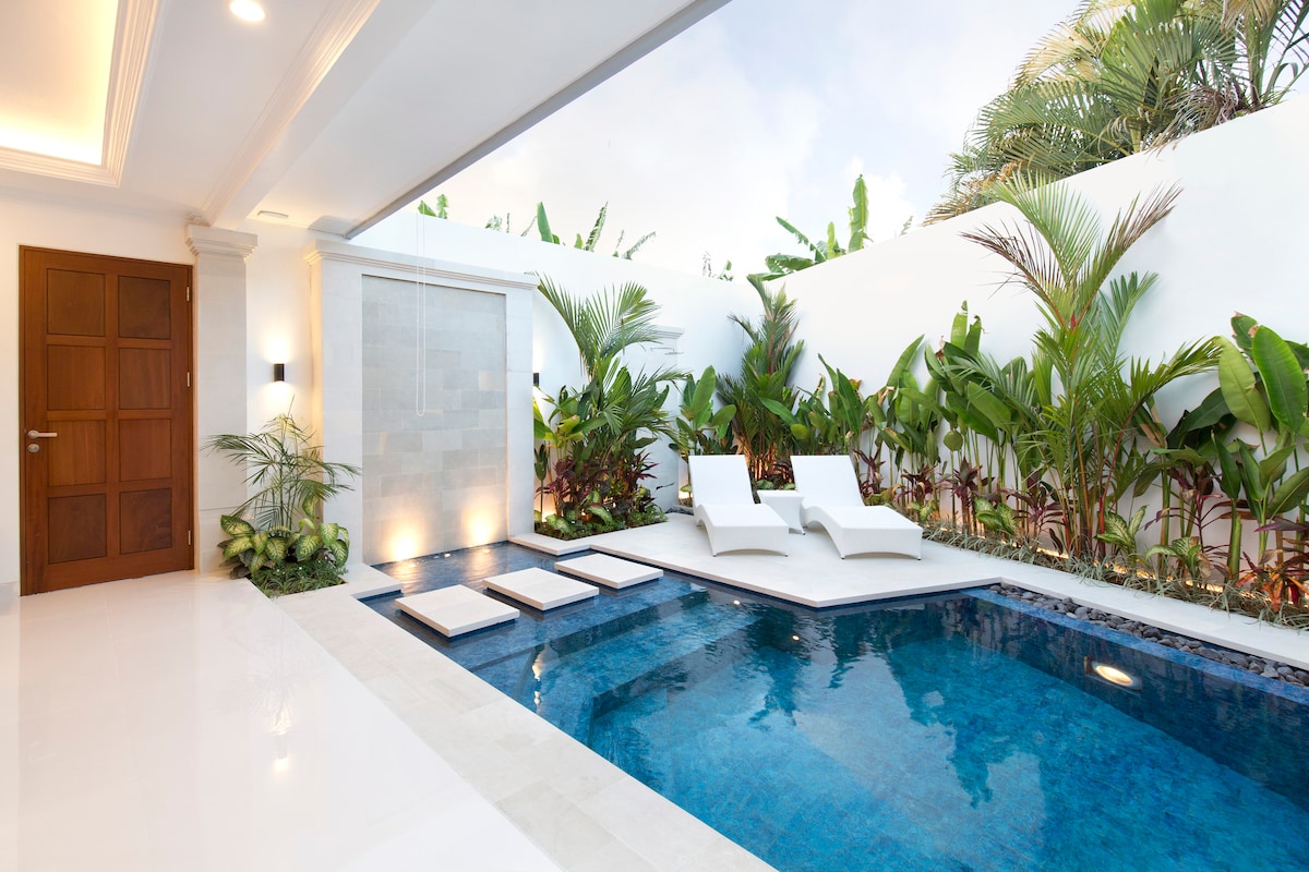 A serene private pool area is surrounded by lush tropical plants, complemented by two sunbeds. The pool features a clean, inviting blue water surface and is designed with a modern aesthetic, creating a peaceful retreat within the villa.