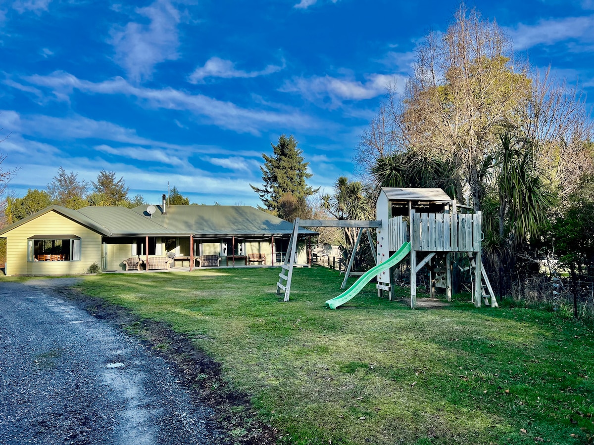 A spacious green lawn surrounds a single-story building, featuring a covered porch along the front. A wooden play structure with a slide is positioned in the yard, creating an inviting play area for families. The sky exhibits a mix of blue and white clouds.