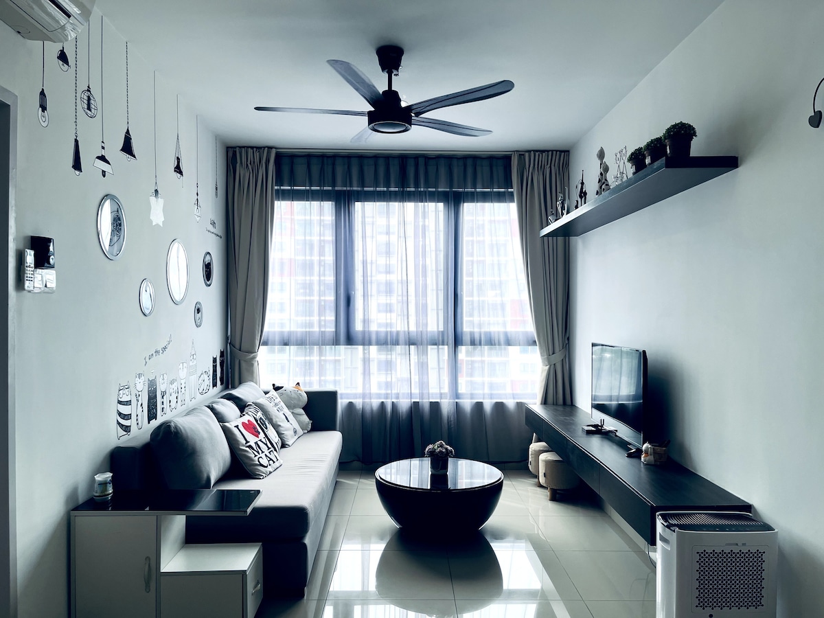 A modern living area is presented, featuring a spacious grey sofa and a sleek circular coffee table. A flat-screen TV is mounted on a low-profile shelf underneath a large window, allowing ample natural light to illuminate the room. Decorative mirrors adorn the wall, adding depth.