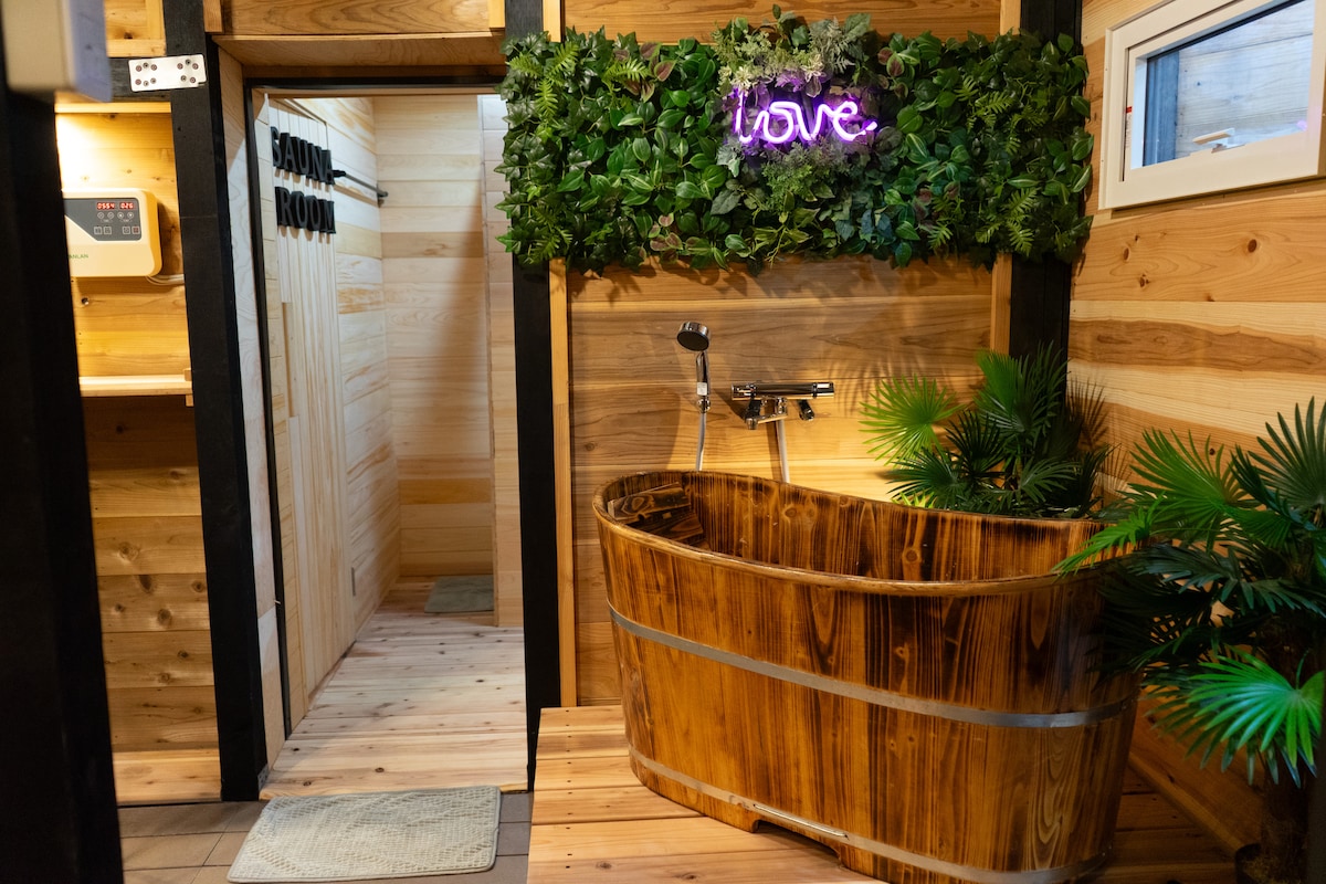 A wooden bathtub is situated in a warmly lit space, featuring natural wood paneling and greenery. A neon sign spelling 'love' adds a modern touch. A door leads to the sauna room, enhancing the serene atmosphere.