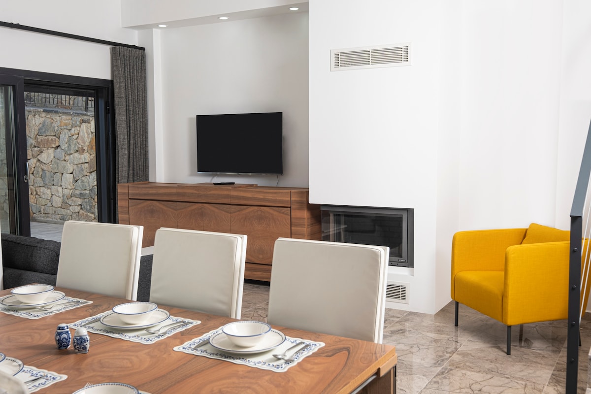 A modern living area is depicted with a wooden dining table set for six, featuring white plates and blue napkins. A sleek, low media console sits opposite a mounted television, while a bright yellow armchair adds a pop of color to the neutral-toned space.