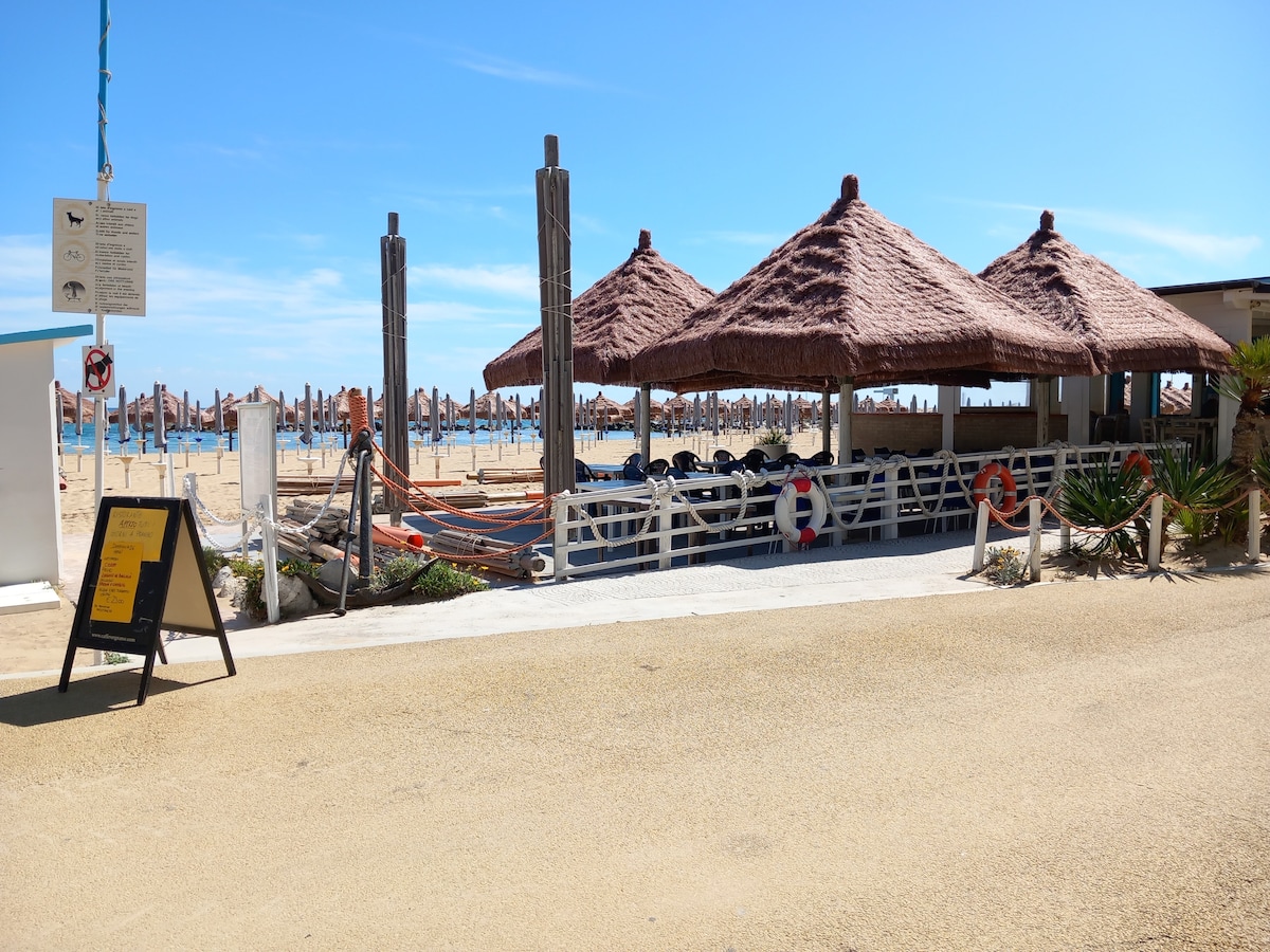 A sandy pathway leads to a beach area featuring thatched-roof huts, providing shaded seating. Wooden posts mark the beach zone, with sun loungers arranged nearby. A signboard stands on the left, and a clear blue sky is visible above, creating a relaxed coastal environment.