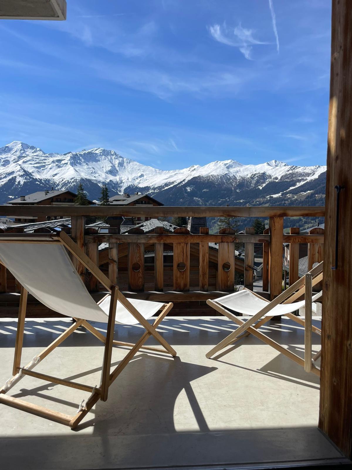 A sunlit balcony is furnished with two wooden lounge chairs, offering a view of the snow-capped mountains in the distance. The wooden railing adds a rustic touch, while a clear blue sky enhances the serene atmosphere of the outdoor space.