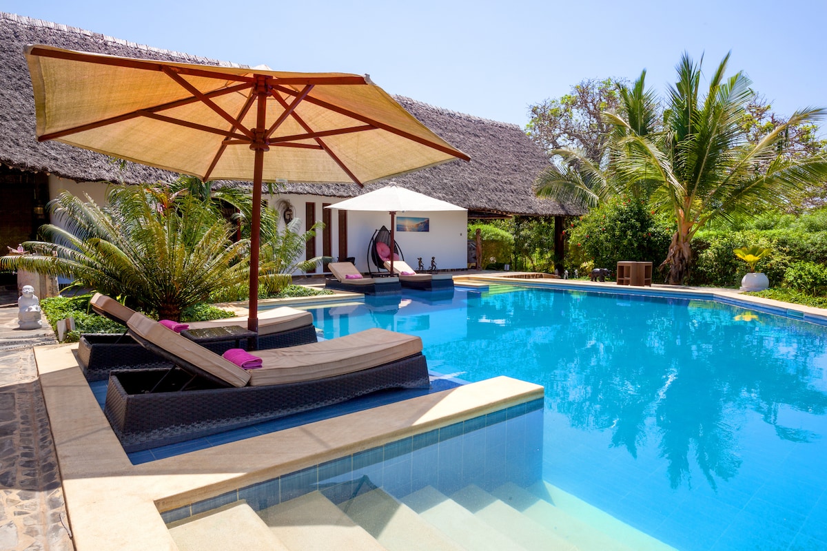 A private pool area is surrounded by lush greenery, featuring comfortable lounge chairs and umbrellas for shade. A modern cabana is present along the edge of the pool, complemented by palm trees and tropical plants enhancing the serene ambiance.