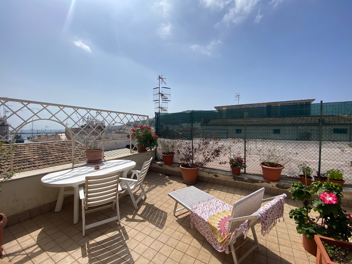 A spacious terrace is presented, featuring a round dining table surrounded by several chairs. A lounge chair with a colorful blanket rests nearby. Potted plants enhance the space, and a partial view of the surrounding area is visible in the background.