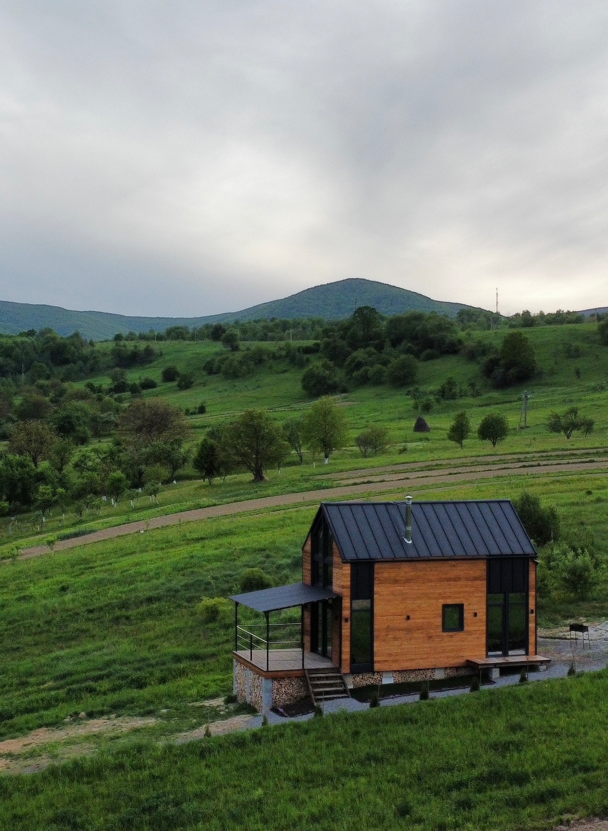 A modern wooden house is set against a backdrop of rolling green hills and mountains. The structure features a black roof and large windows, allowing for a connection with the surrounding landscape. A small porch extends from the front, inviting outdoor relaxation.