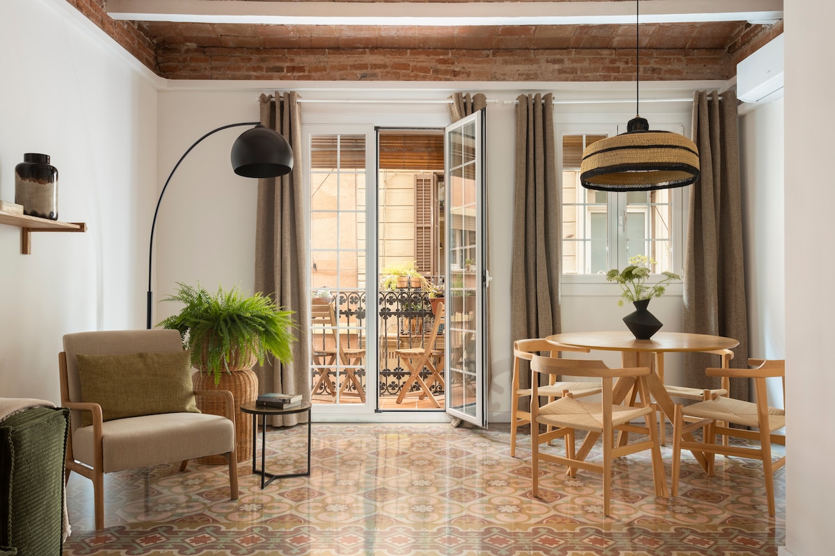 A spacious living area is highlighted by large glass doors that open to a balcony, allowing natural light to fill the room. A round dining table, surrounded by four chairs, is complemented by a stylish floor lamp. Decorative plants and a textured rug add warmth to the space.